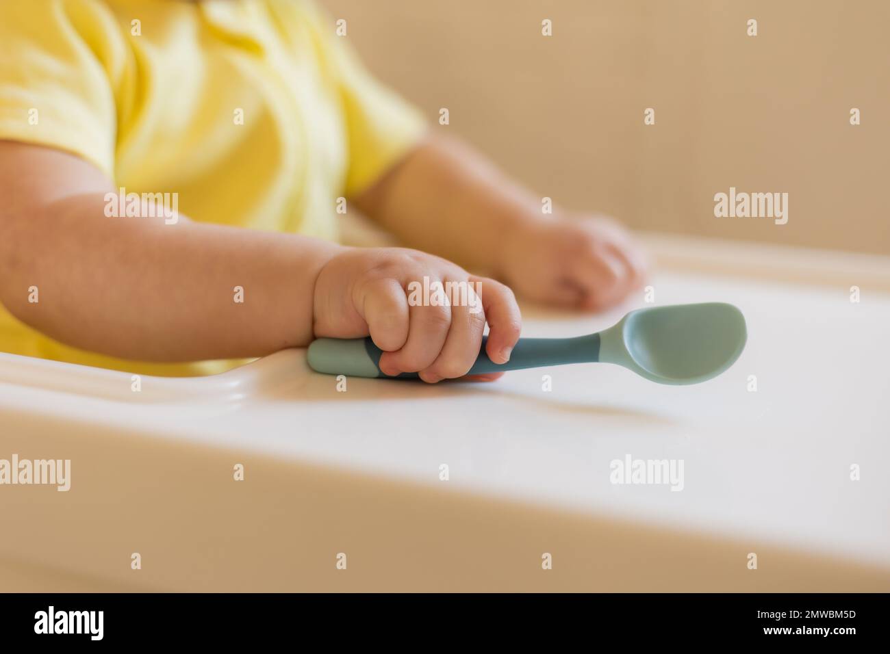 un bébé mignon dans un body jaune est assis dans une chaise haute dans la cuisine de la maison avec une cuillère en plastique. Nourriture pour bébés, première nourriture complémentaire. Hin Banque D'Images