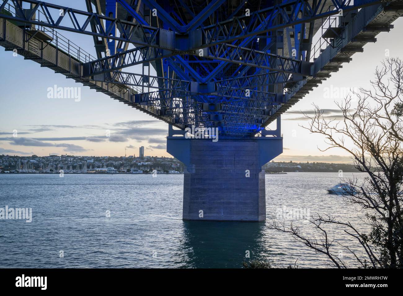 Piliers en béton et poutres en acier sous le pont du port d'Auckland illuminés de lumière bleue. Un bateau-ferry qui se dirige vers le pont. Banque D'Images