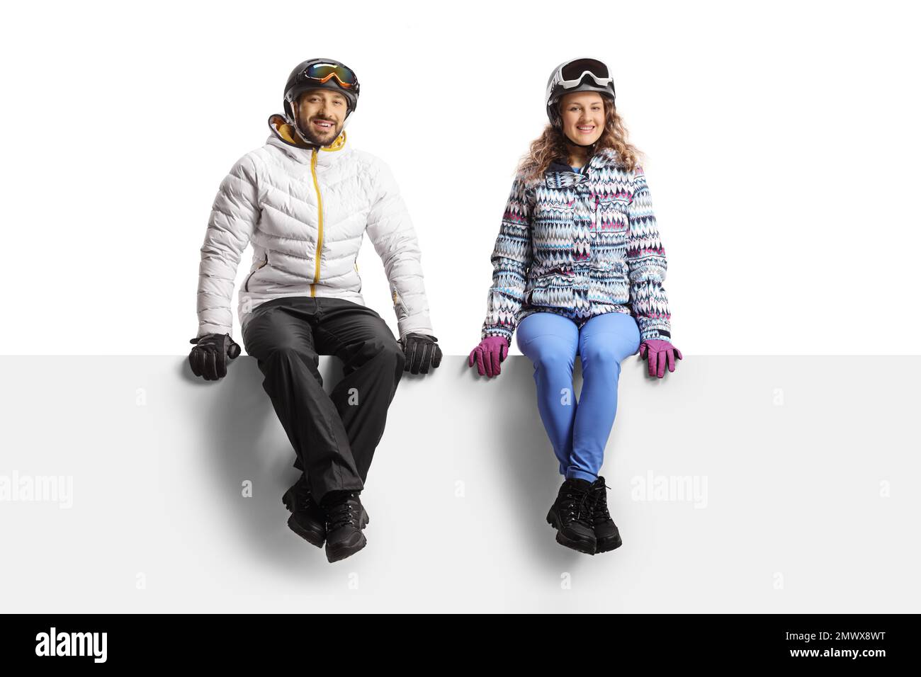 Jeune homme et jeune femme portant des vestes et des casques d'hiver et assis sur un panneau vierge isolé sur fond blanc Banque D'Images