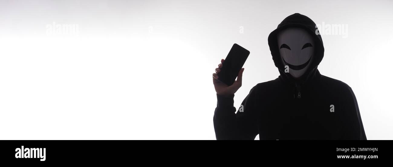 Hacker Anonyme et masque de visage avec smartphone en main. Homme en chemise noire à capuche tenant et utilisant un téléphone portable sur fond blanc. Représentent les CR cybernétiques Banque D'Images