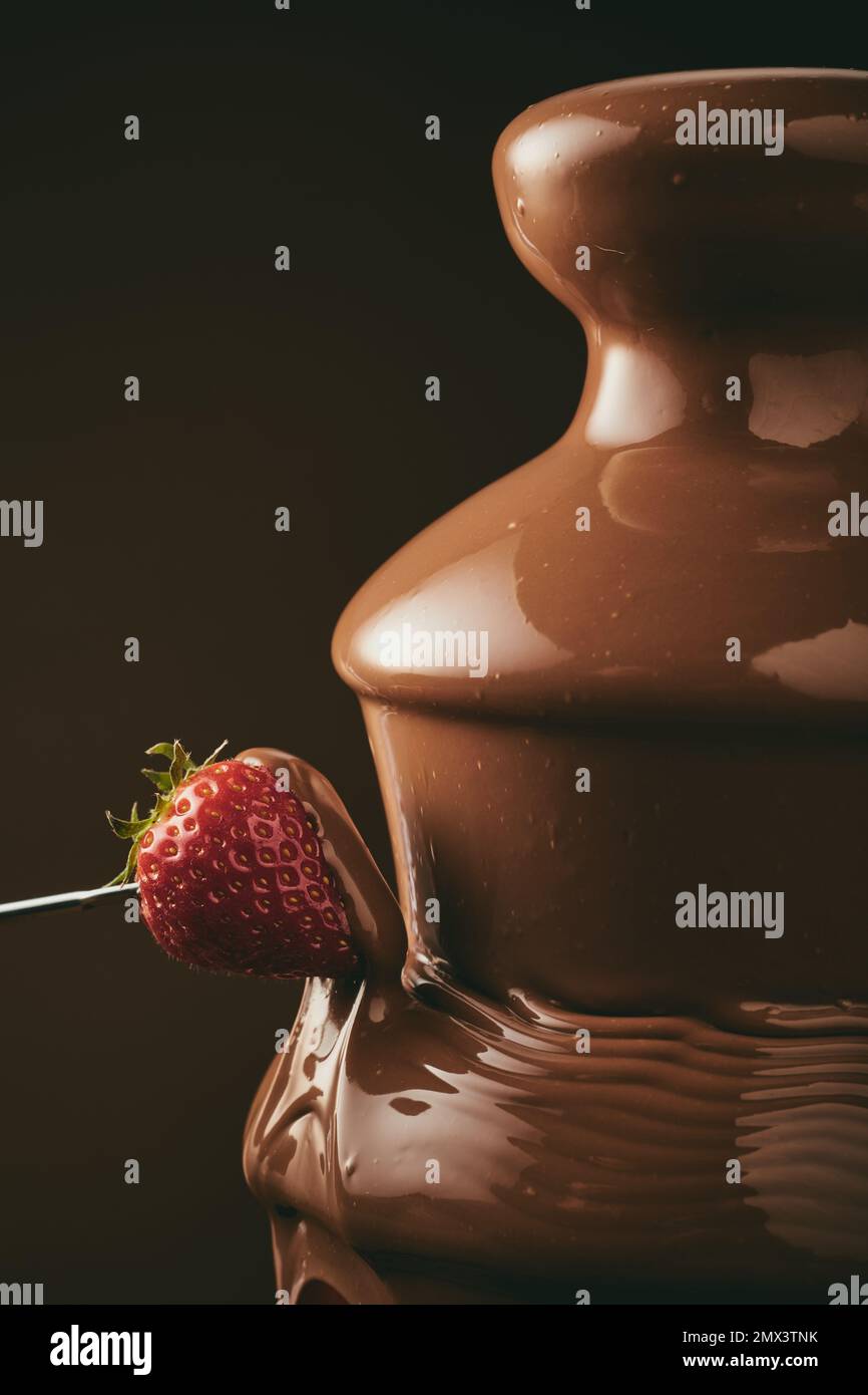 Personne anonyme en trempant de la fraise fraîche sur un cure-dent dans une fontaine de chocolat doux pendant la fête sur fond noir Banque D'Images