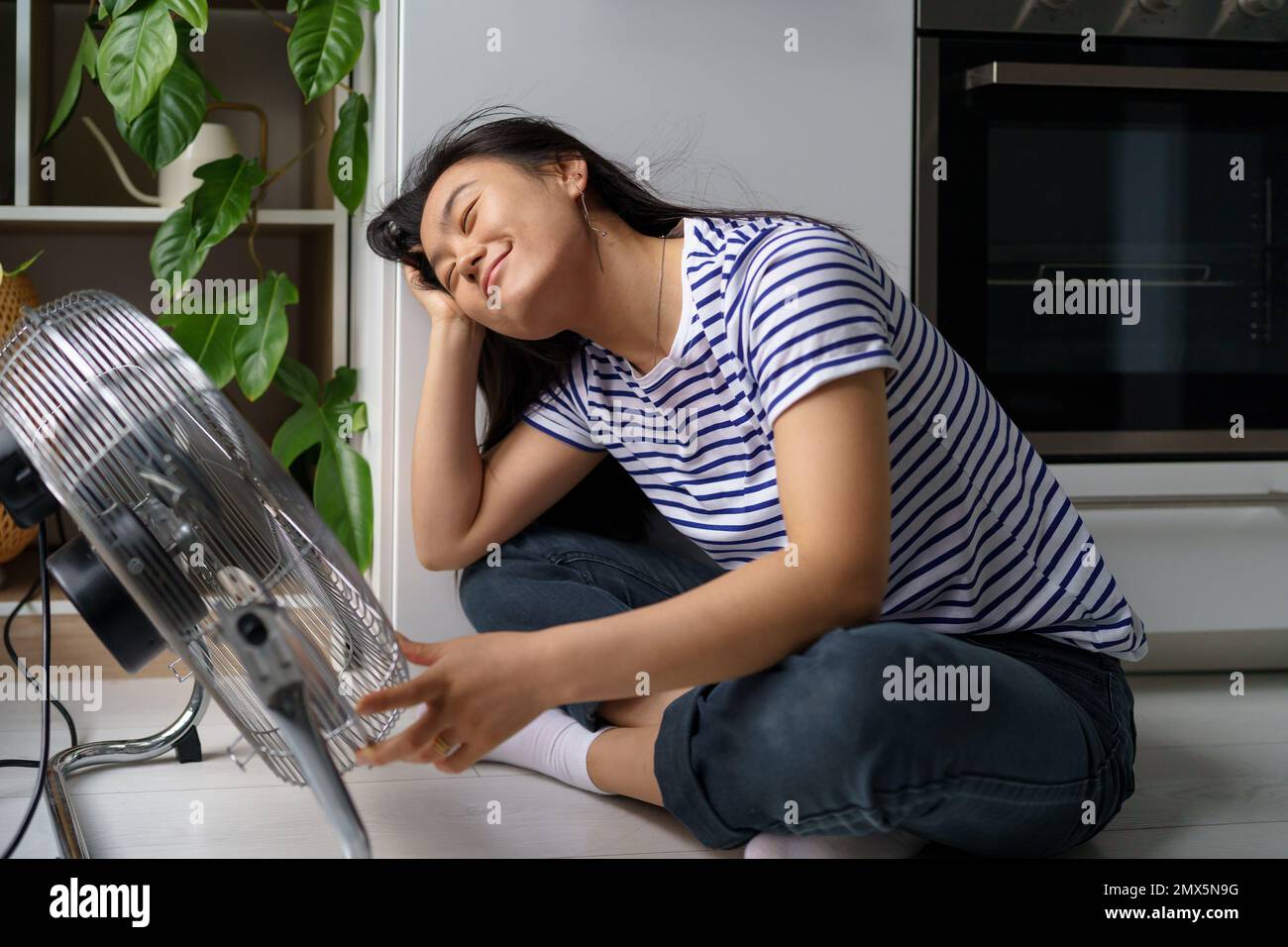 Heureuse fille asiatique détendue se réjouit dans le flux d'air froid du ventilateur se trouve sur le sol dans la cuisine de l'appartement Banque D'Images