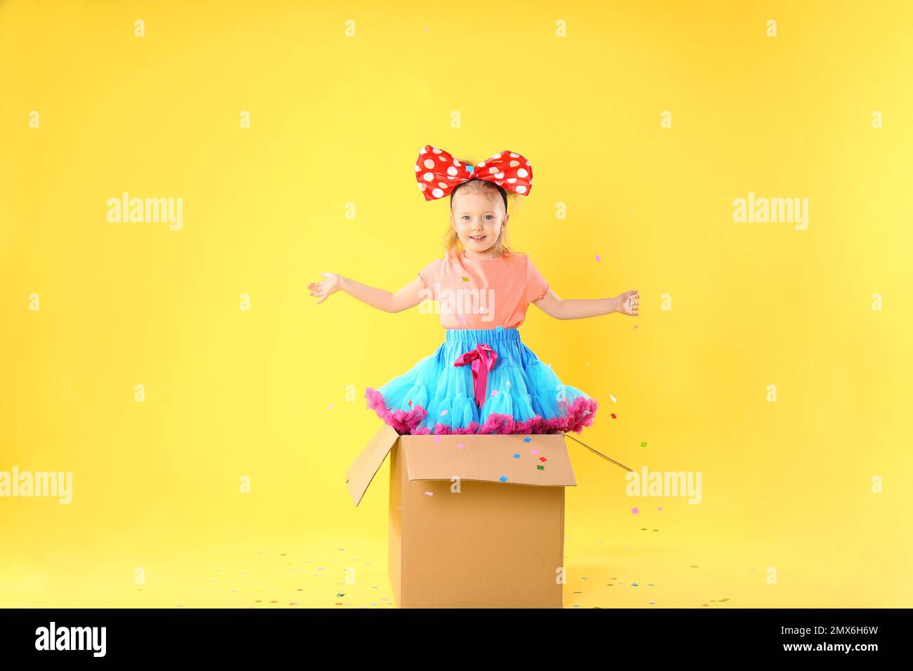 Petite fille avec grand noeud dans boîte en carton sous douche confetti sur fond jaune. Le jour de l'imbécile d'avril Banque D'Images