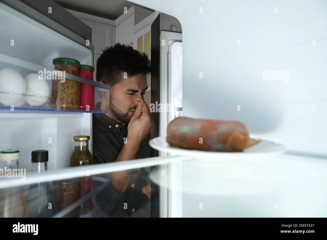 Jeune homme sentant une mauvaise odeur de saucisse gâtée dans le réfrigérateur, vue de l'intérieur Banque D'Images