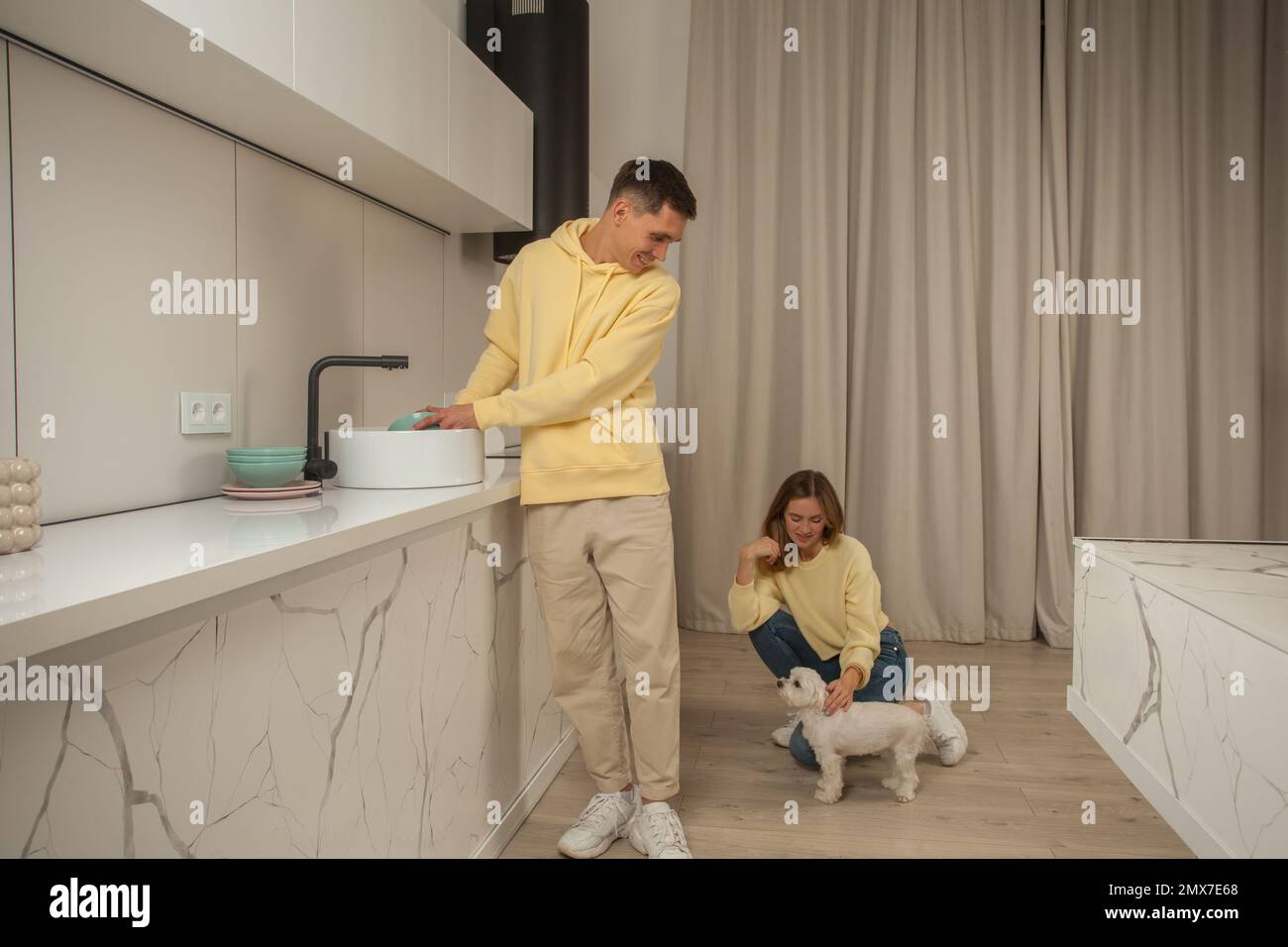 Homme lavant les plats dans l'évier et femme assis et petting chien blanc Banque D'Images