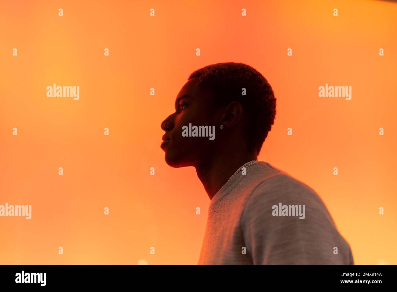 Vue latérale d'un homme afro-américain pensif dans un chandail décontracté debout contre la lumière orange néon et regardant loin avec soin en studio Banque D'Images