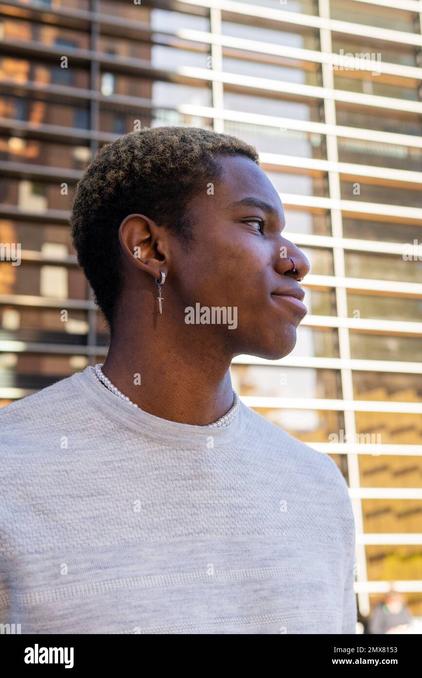 Vue latérale d'un homme afro-américain confiant dans des vêtements décontractés avec boucles d'oreilles en regardant loin tout en se tenant près du mur du bâtiment Banque D'Images