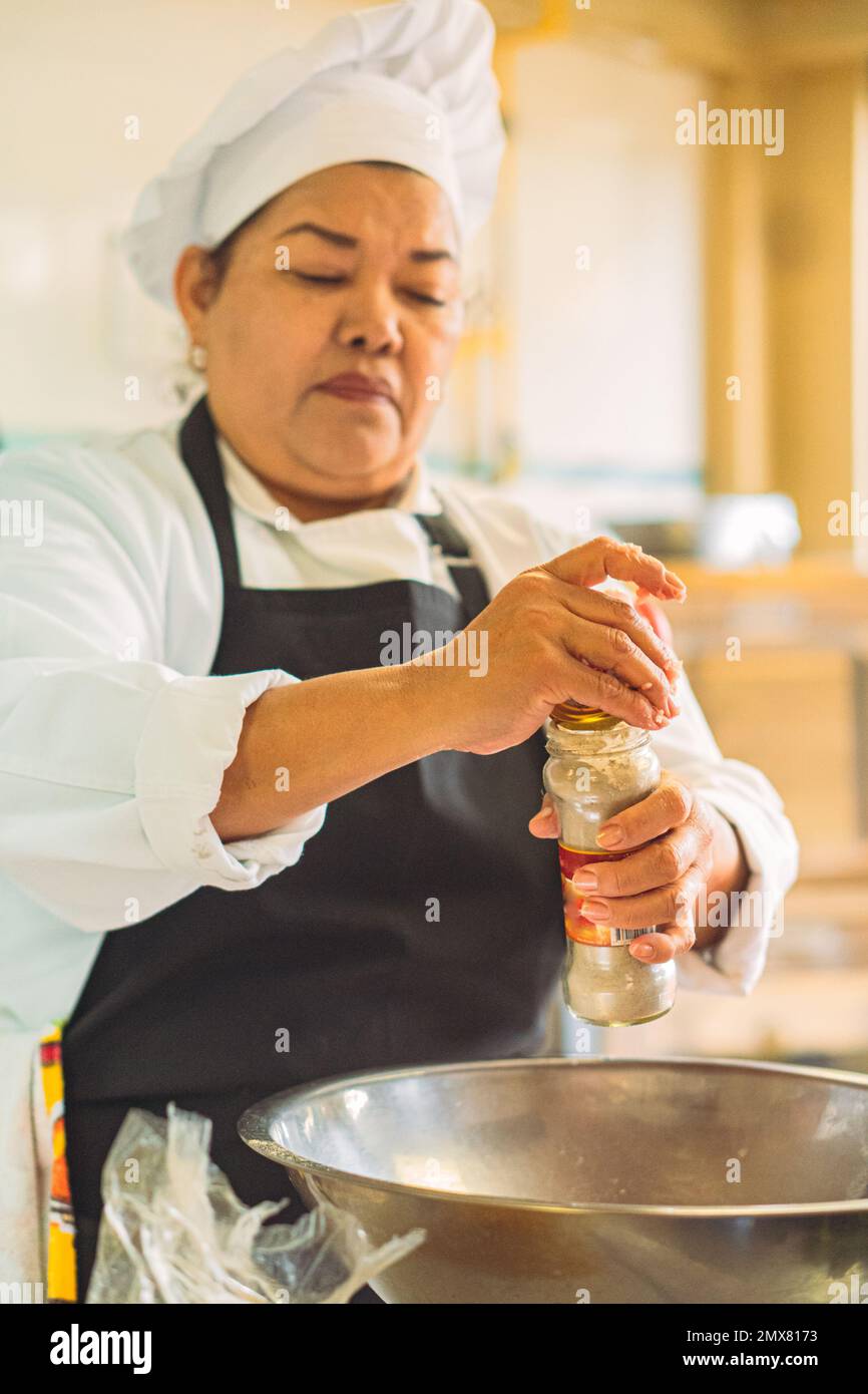 Chef féminin concentré en uniforme et en tablier ajoutant des épices dans un bol en métal tout en préparant la nourriture dans la cuisine du restaurant Banque D'Images