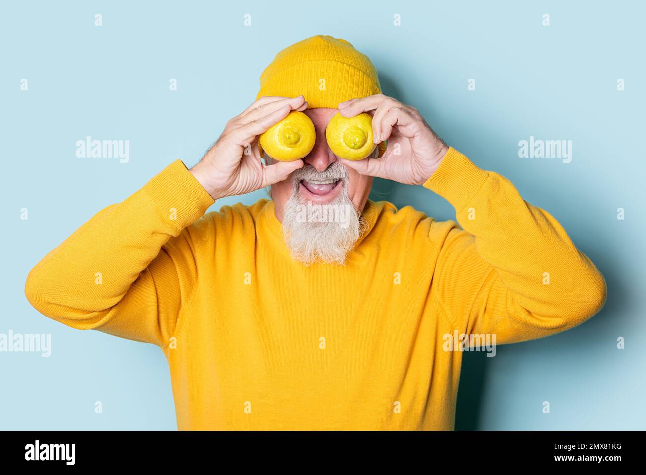 Surprise de mâle âgé barbu dans un chandail jaune et un chapeau couvrant les yeux avec des citrons sur fond bleu Banque D'Images