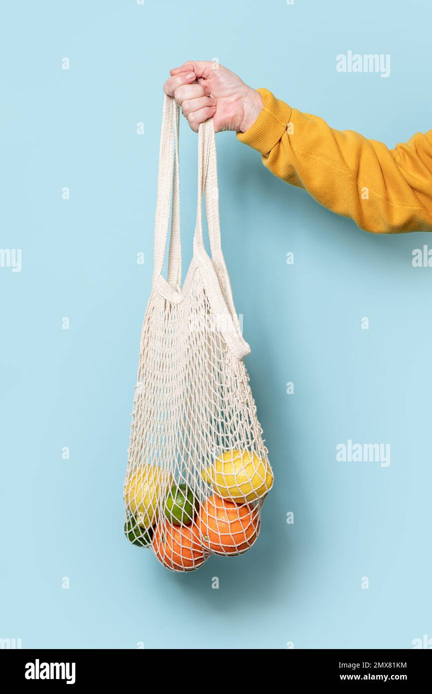 Vue latérale d'une petite femme sénior non reconnaissable dans un chandail jaune tenant un sac en filet avec des fruits sur fond bleu Banque D'Images