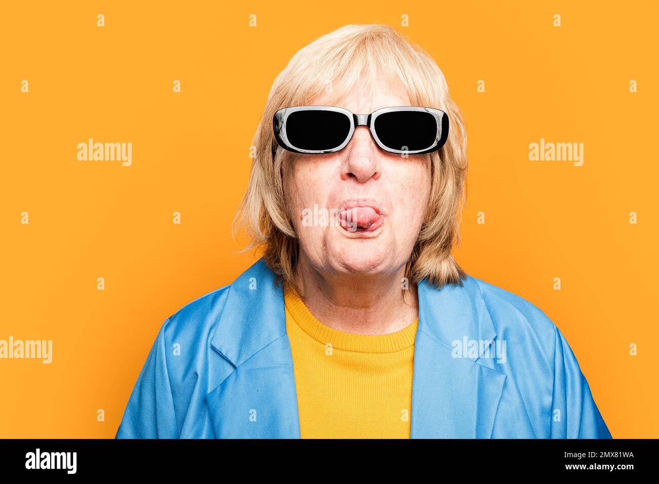 Portrait d'une femme âgée dans des lunettes de soleil tendance et des vêtements brillants regardant l'appareil photo tout en montrant la langue sur fond orange Banque D'Images