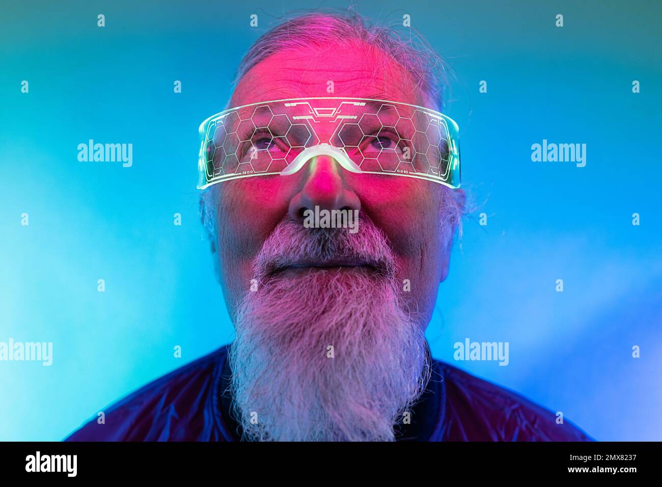 Portrait d'un hipster mâle barbu âgé sérieux dans des verres regardant vers le haut tout en se tenant sur fond bleu éclairé par une lumière de néon Banque D'Images