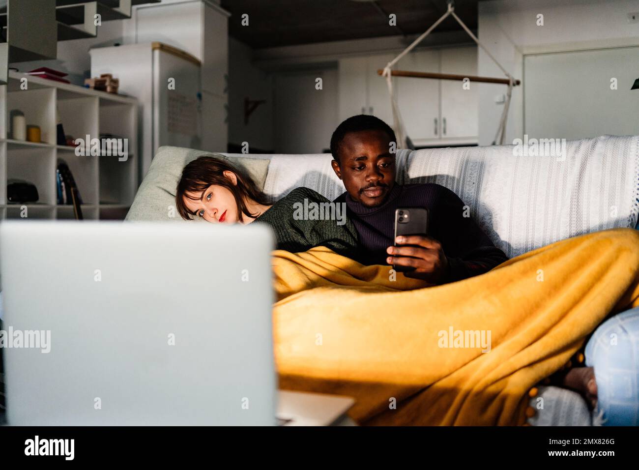Un homme afro-américain concentré vérifie les médias sociaux tout en se reposant sur un canapé sous une couverture avec une petite amie regardant un film sur un ordinateur portable dans le salon Banque D'Images