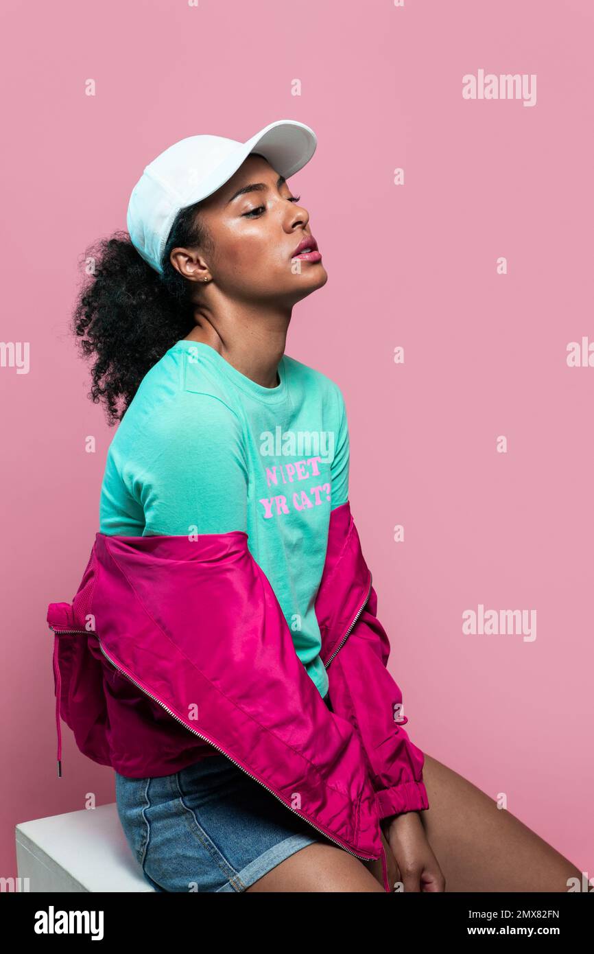 Jeune femme afro-américaine confiante en tenue décontractée et casquette brillante assise sur une chaise et regardant loin sur fond rose Banque D'Images