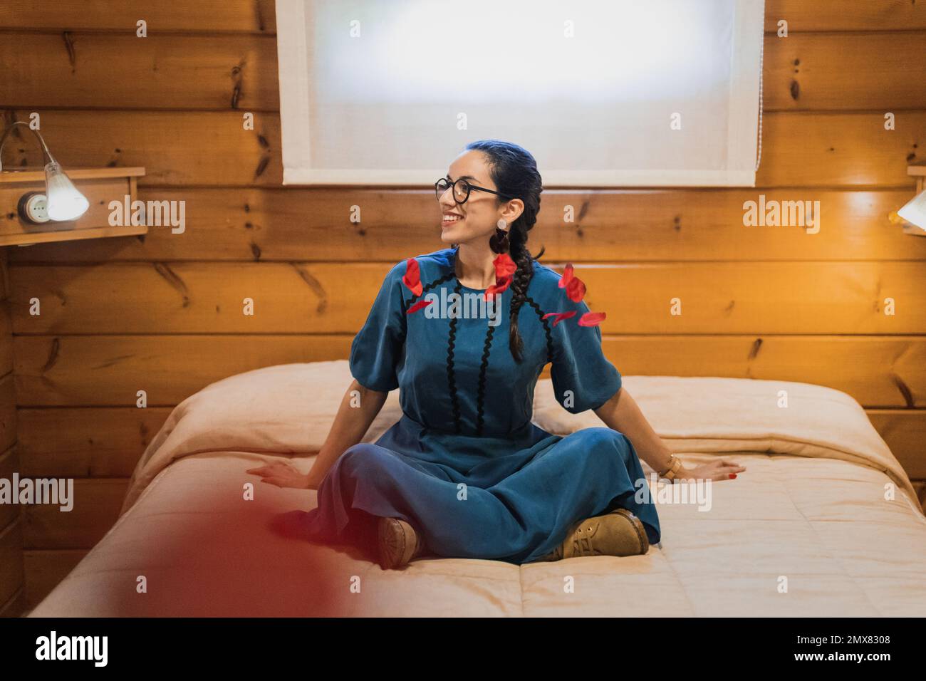 Corps complet de jeune femme optimiste en lunettes assis sur le lit et souriant tout en jetant des pétales pendant le week-end Banque D'Images