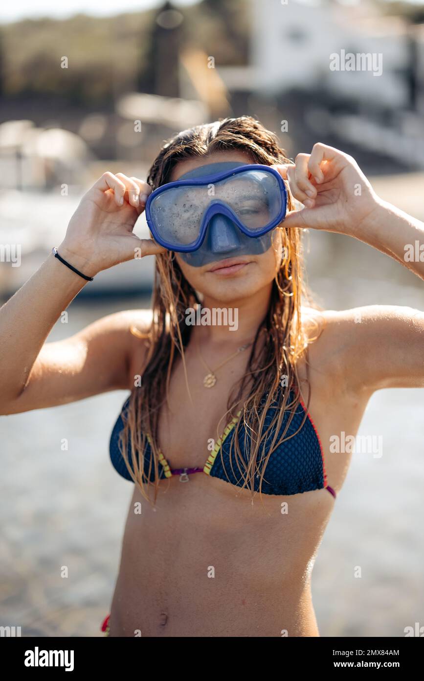Slim femme plongeur en maillot de bain et lunettes pour plonger sourire et regarder l'appareil photo tout en se relaxant le week-end d'été sur la plage de Cadaques dans le dos éclairé Banque D'Images