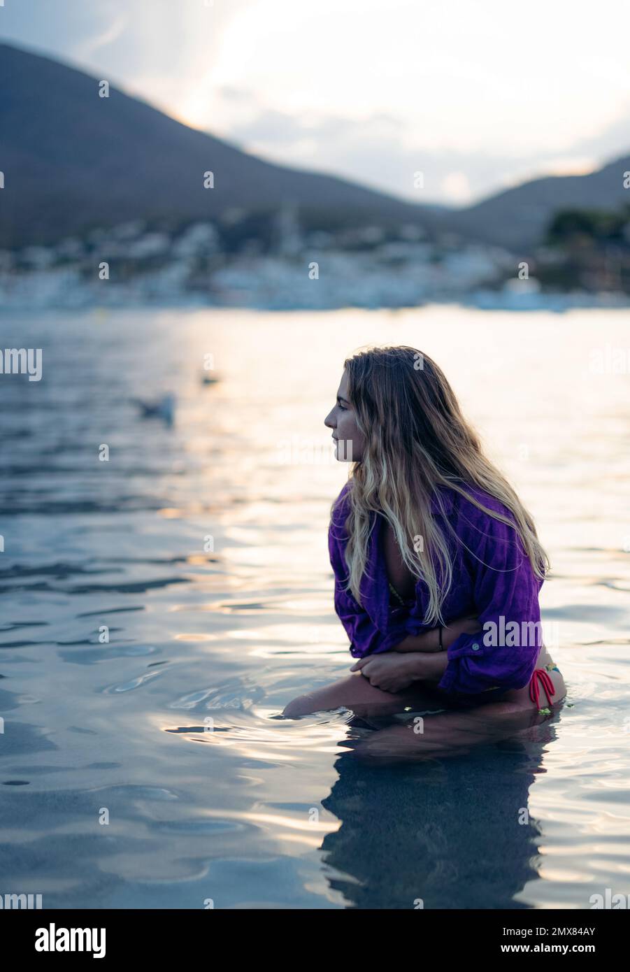 Jeune femme rêveuse avec de longs cheveux blonds dans le bikini et chemise pourpre assis dans les eaux peu profondes de la mer et regardant loin sur la plage de Cadaques au coucher du soleil Banque D'Images