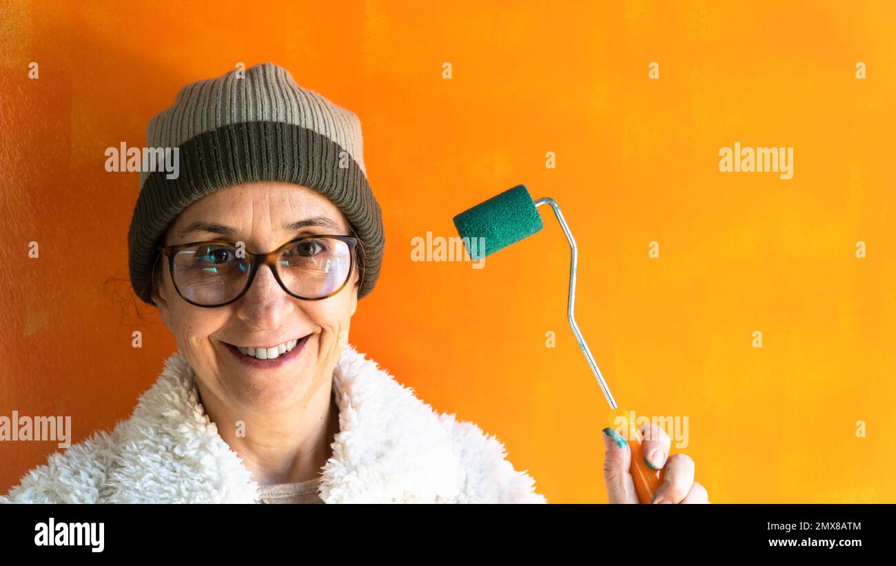 Femme d'âge moyen peint un mur. Portrait d'une femme souriante tenant une brosse rotative. Déménagement. Mise au point sélective incluse. Bruit et grain inclus Banque D'Images