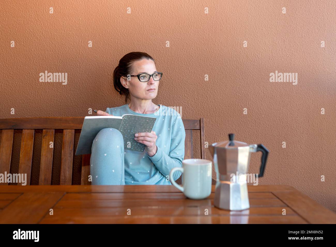 Femme assise dans son pyjama et essorée dans un carnet, café du matin sur une table en bois. Banque D'Images