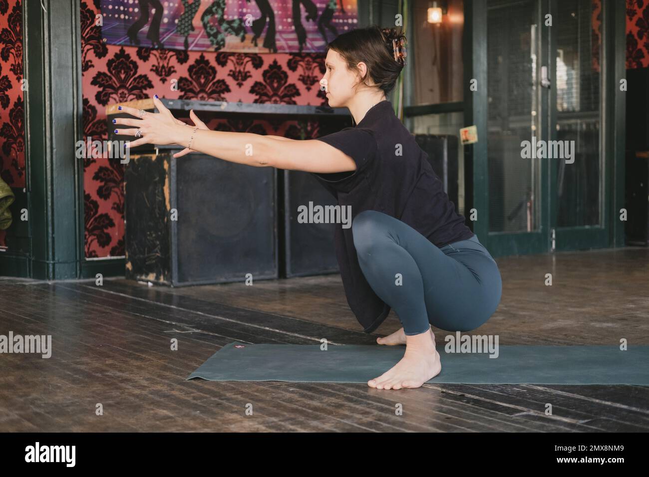 Femme yogi nouvelle enseignante en malasana, squat ou guirlande pose pendant son entraînement de yoga, pieds nus et portant des vêtements de sport verts et noirs sur un tapis sur le corps Banque D'Images