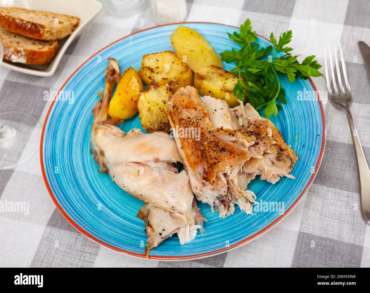 Filet de lapin rôti et jambe sur une assiette bleue avec pommes de terre cuites au four servi avec un verre de vin sur une table en bois dans une cafétéria Banque D'Images