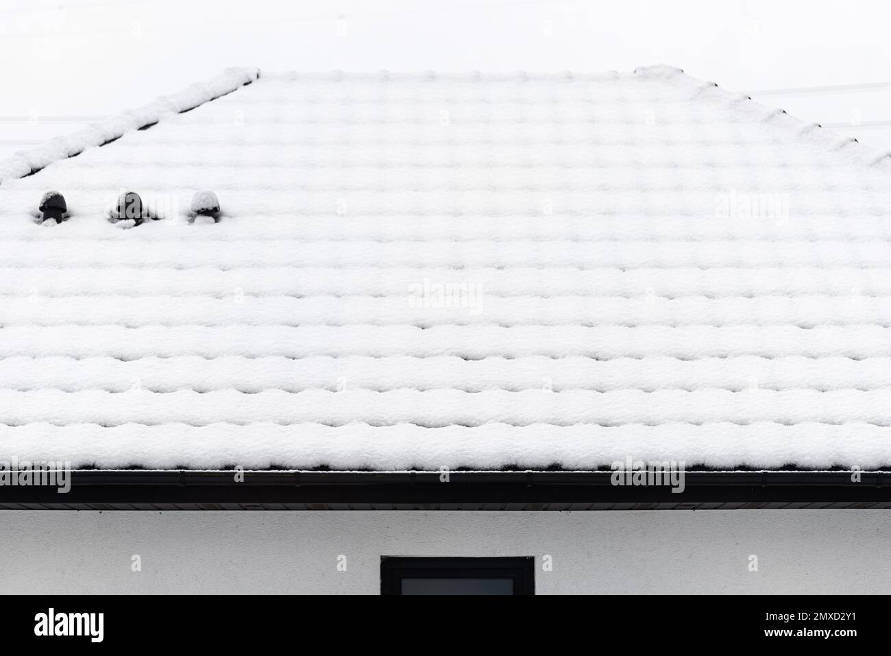 Le toit d'une maison unifamiliale est couvert de neige sur un ciel nuageux, d'une cheminée de ventilation en céramique visible sur le toit et de neige tombant. Banque D'Images