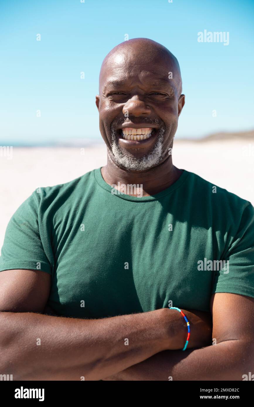 Homme senior afro-américain sans souci et chauve-souris avec des armes croisées souriant à la plage contre un ciel dégagé Banque D'Images