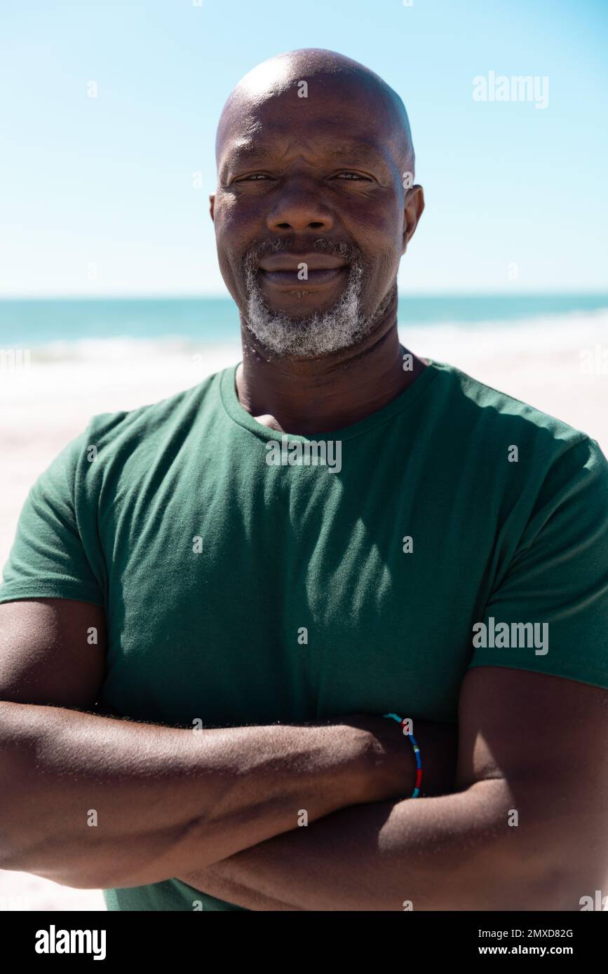 Portrait d'un homme âgé afro-américain confiant et chauve avec des armes croisées à la plage sous un ciel dégagé Banque D'Images