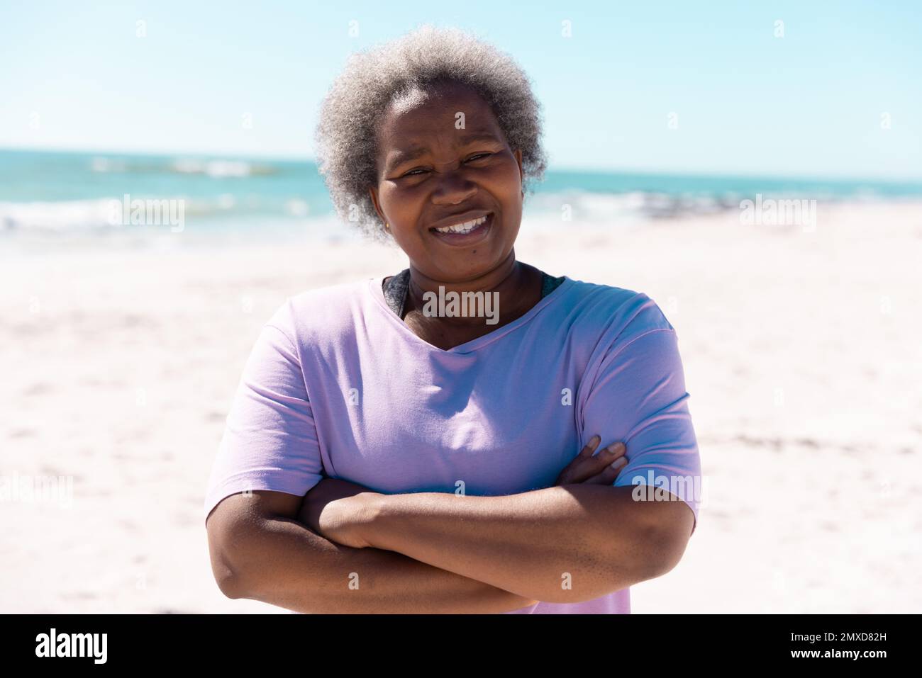 Femme afro-américaine souriante aux bras croisés debout à la plage sous ciel clair en été Banque D'Images