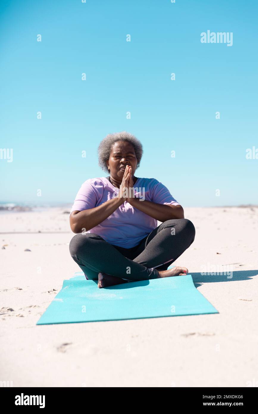 Femme afro-américaine âgée à pattes croisées méditant sur un tapis à la plage contre un ciel bleu clair Banque D'Images