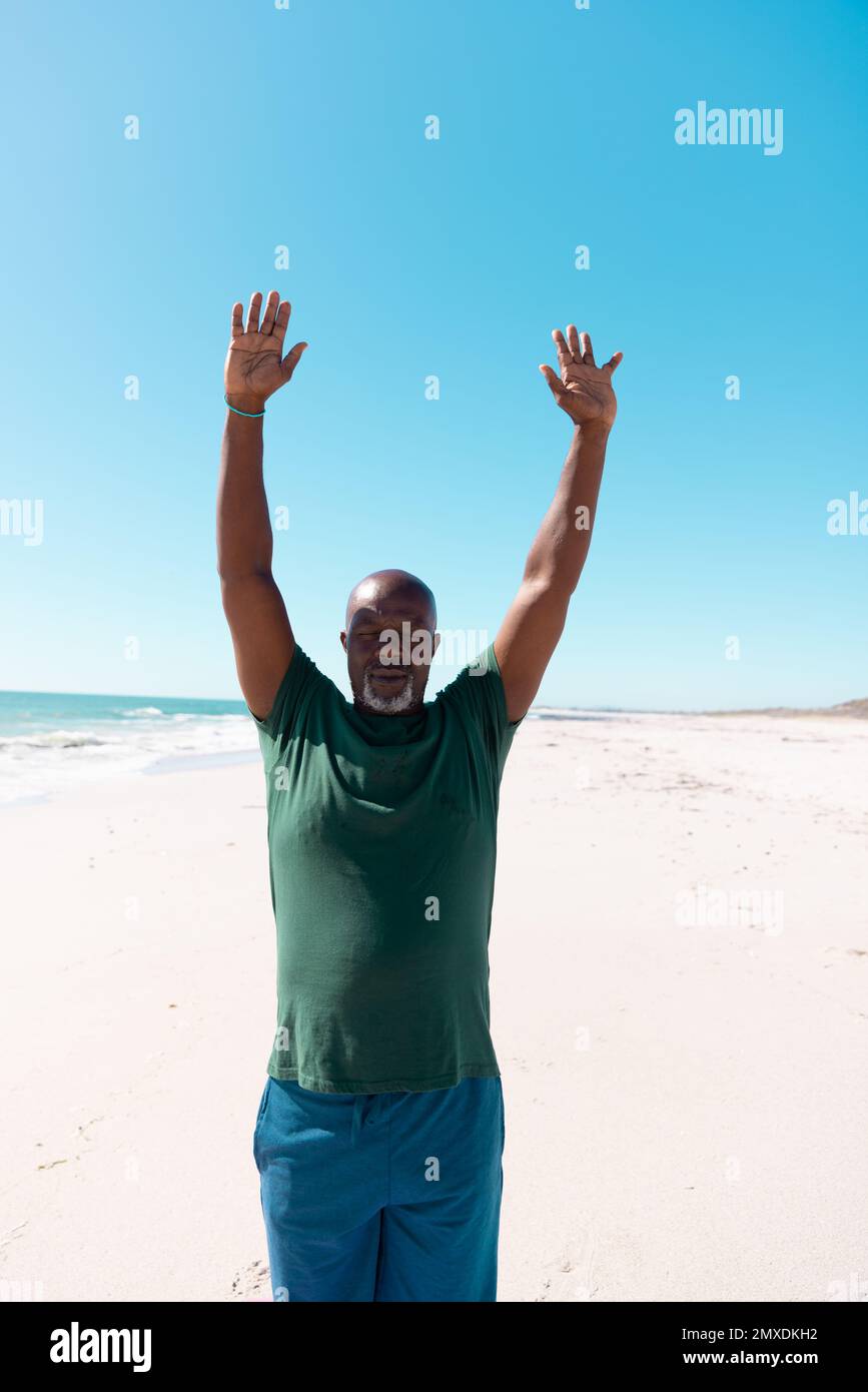 Homme âgé chauve d'Amérique d'Afrique avec des armes levées méditant sur la plage de sable sous ciel clair Banque D'Images