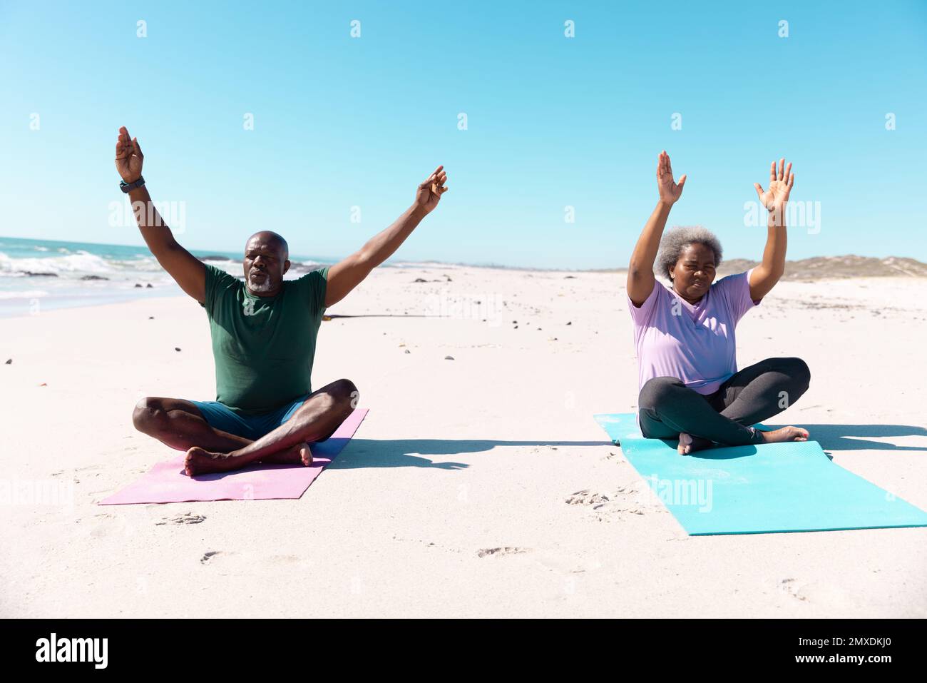 Couple afro-américain âgé avec des armes levées méditant tout en étant assis sur des tapis à la plage sous le ciel Banque D'Images