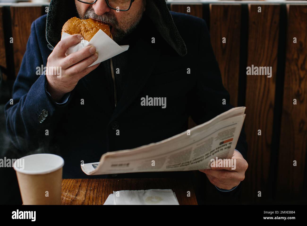 Un barbu d'âge moyen mange un savoureux hamburger et lit le journal d'aujourd'hui dans une rue de la ville en hiver. Concept de cuisine de rue. Banque D'Images