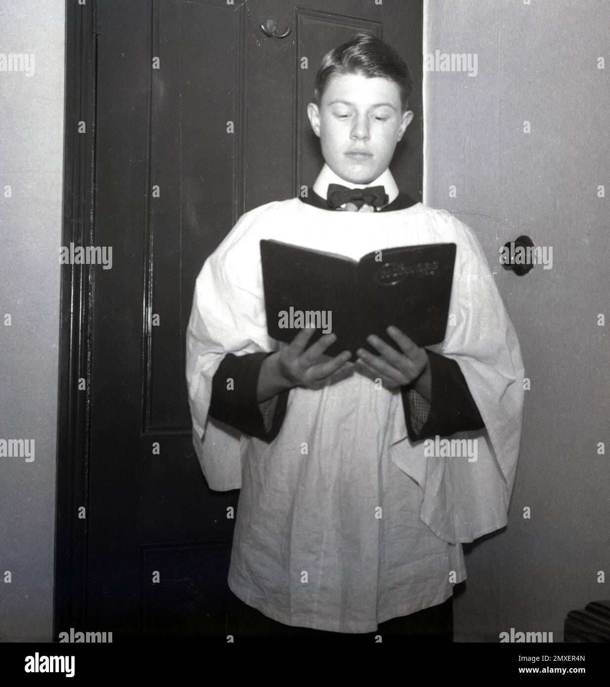 1950s, historique, un choirboy adolescent debout à l'intérieur d'une porte portant la robe traditionnelle ou le cassock et tenant un livre de chant, Angleterre, Royaume-Uni. Banque D'Images