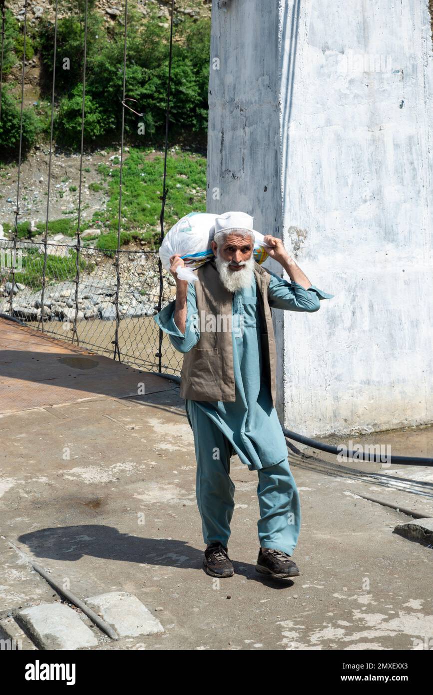 Vieil homme à la barbe blanche portant un sac blanc sur le pont suspendu du village de Mankar, vallée de Swat, Pakistan Banque D'Images
