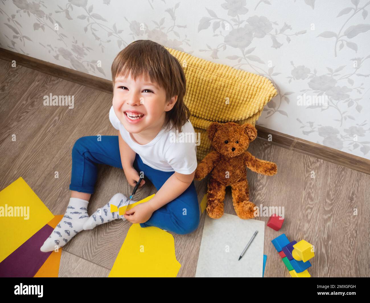 Le jeune garçon apprend à couper du papier de couleur avec des ciseaux. Les enfants sont assis au sol dans la chambre des enfants avec des blocs de jouets et un ours en peluche. Cours d'éducation pour les enfants Banque D'Images