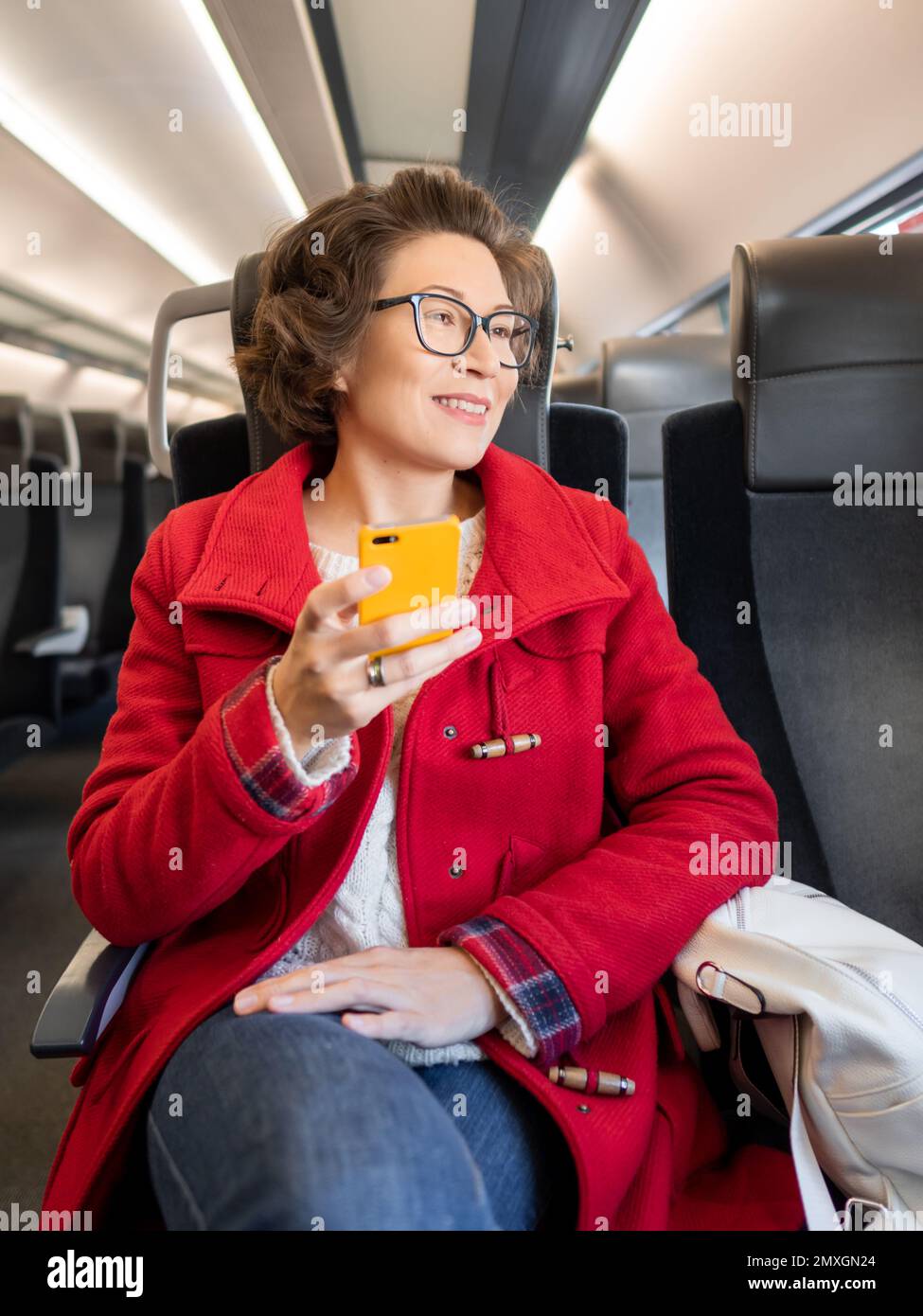 Femme souriante dans un duffle-coat rouge envoyant un SMS sur un smartphone dans un train de banlieue. Voyager en véhicule terrestre. Banque D'Images