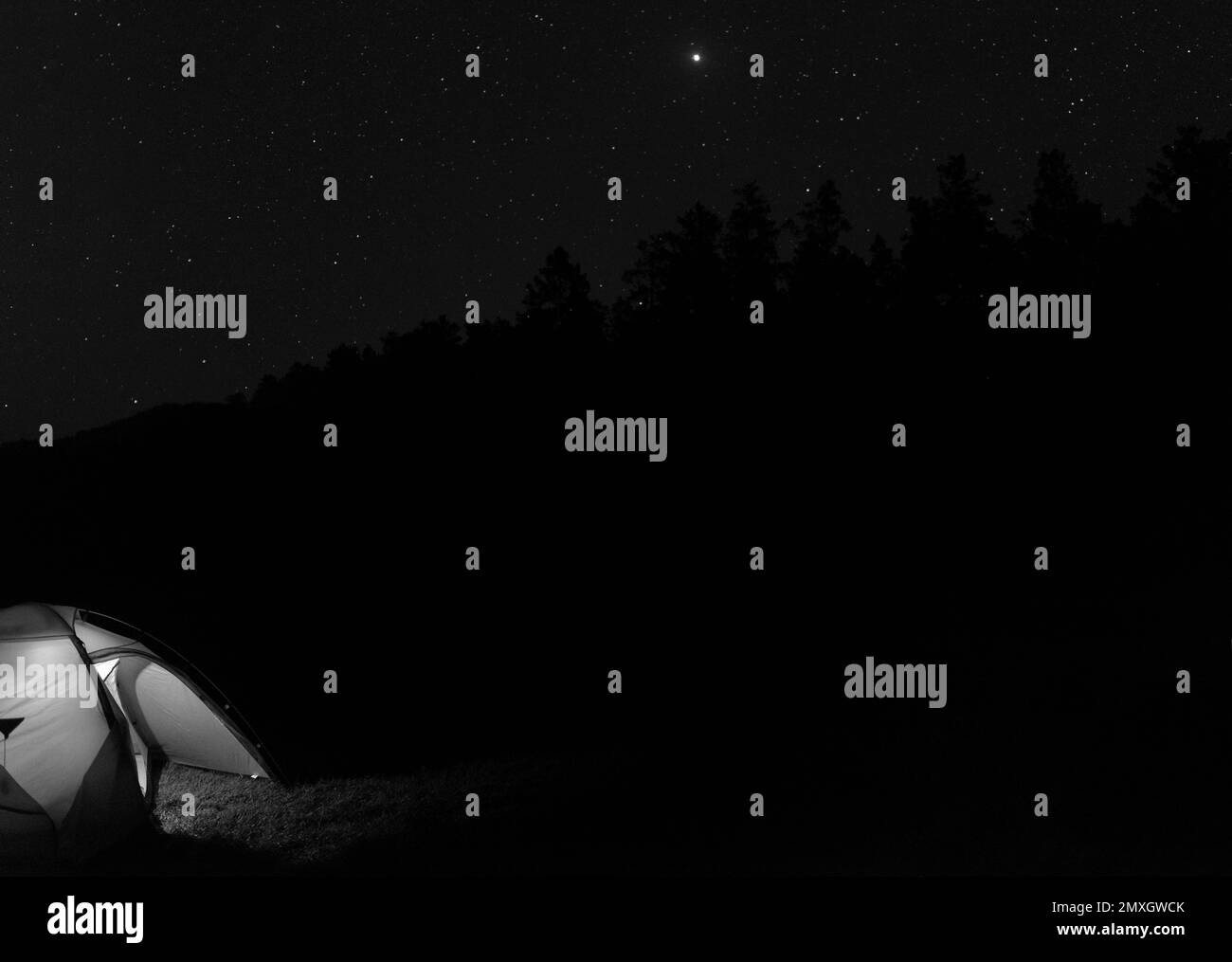 Photo en noir et blanc d'une tente de voyageurs illuminée par une entrée ouverte dans l'obscurité la nuit, avec des étoiles à côté des montagnes et des arbres. Banque D'Images