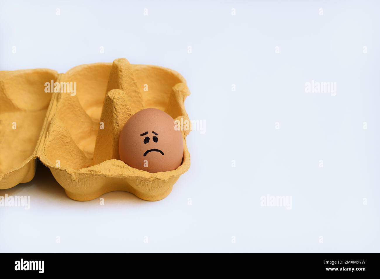Un seul œuf de poulet au visage triste dans un carton jaune ouvert, une demi-douzaine de cartons d'œufs, à gauche du cadre sur une surface blanche et propre Banque D'Images