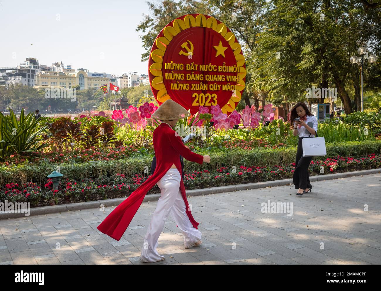 Une femme habillée de la traditionnelle ao dai avec un chapeau conique regarde son smartphone comme une femme habillée à bon prix marche dans la direction opposée au passé Banque D'Images
