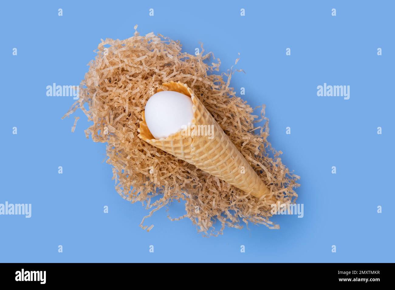 Cornet de crème glacée gaufrée avec un œuf de Pâques blanc caché dans un nid en papier sur fond bleu, concept, minimalisme. Carte de Pâques créative Banque D'Images