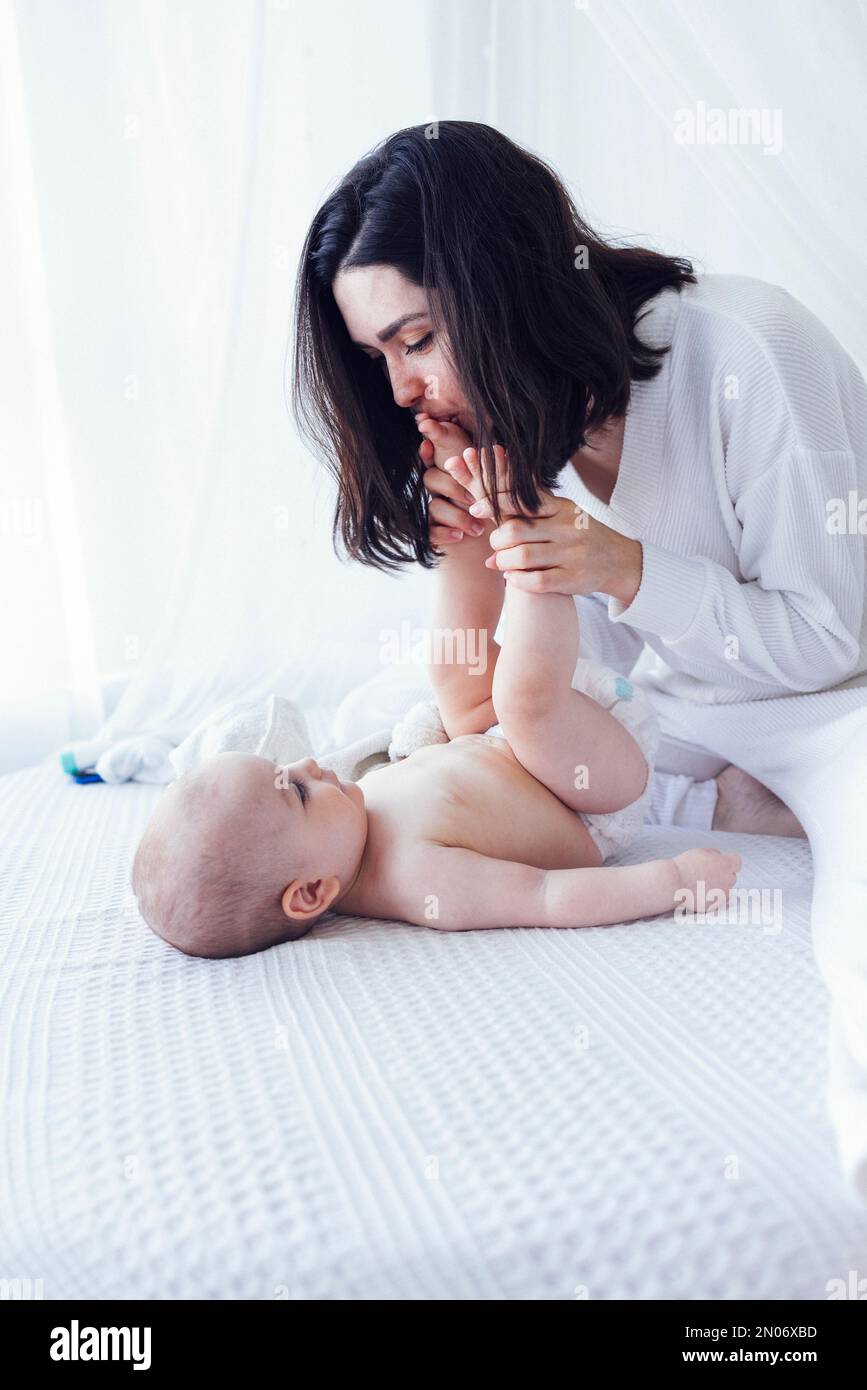 Jeune belle mère embrassant son bébé mignon sur la joue. La brune blanche touche son enfant souriant avec son visage. Joli bébé dans un body blanc avec Banque D'Images