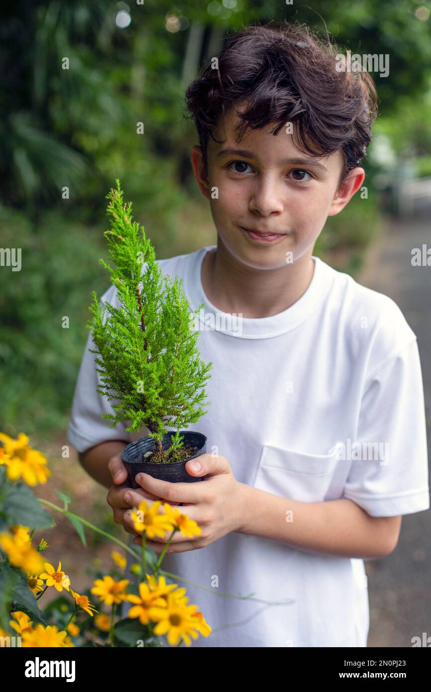 Un garçon tenant un petit arbre dans un pot, debout dans un jardin. Banque D'Images