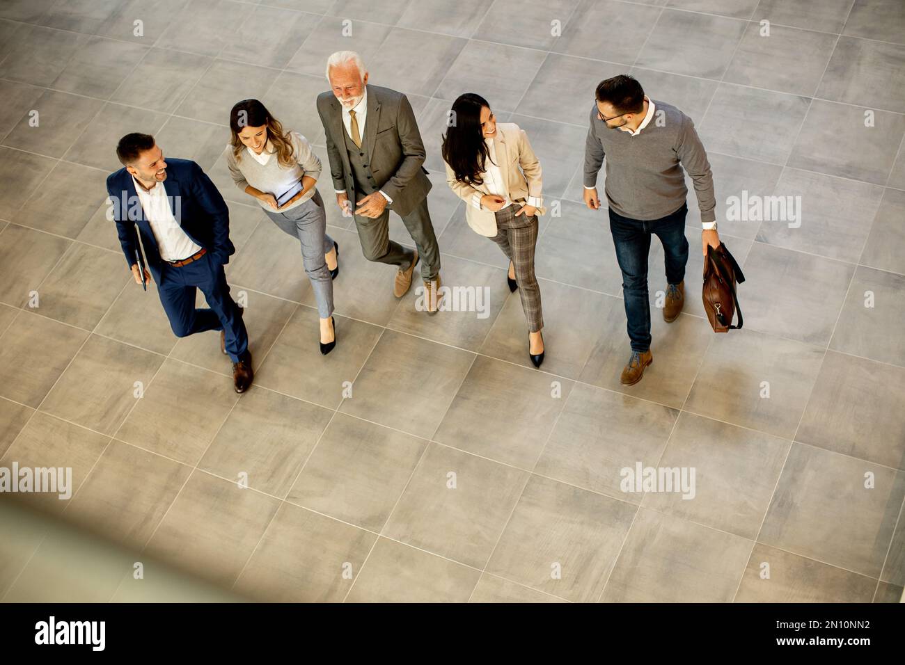 Un groupe de jeunes et de personnes âgées en voyage d'affaires se prompe dans un couloir de bureau, capturé dans une vue aérienne. Ils sont vêtus d'une tenue habillée, ils marchent avec humour Banque D'Images