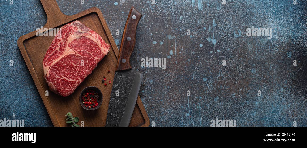 Bœuf à la viande cru bœuf marbré steak de première qualité Ribeye sur planche à découper en bois et fond de table de cuisine rustique en béton d'en haut avec un gros couteau et des épices Banque D'Images