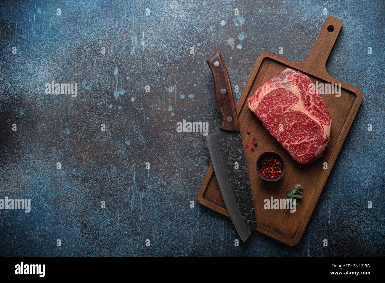 Bœuf à la viande cru bœuf marbré steak de première qualité Ribeye sur planche à découper en bois et fond de table de cuisine rustique en béton d'en haut avec un gros couteau et des épices Banque D'Images