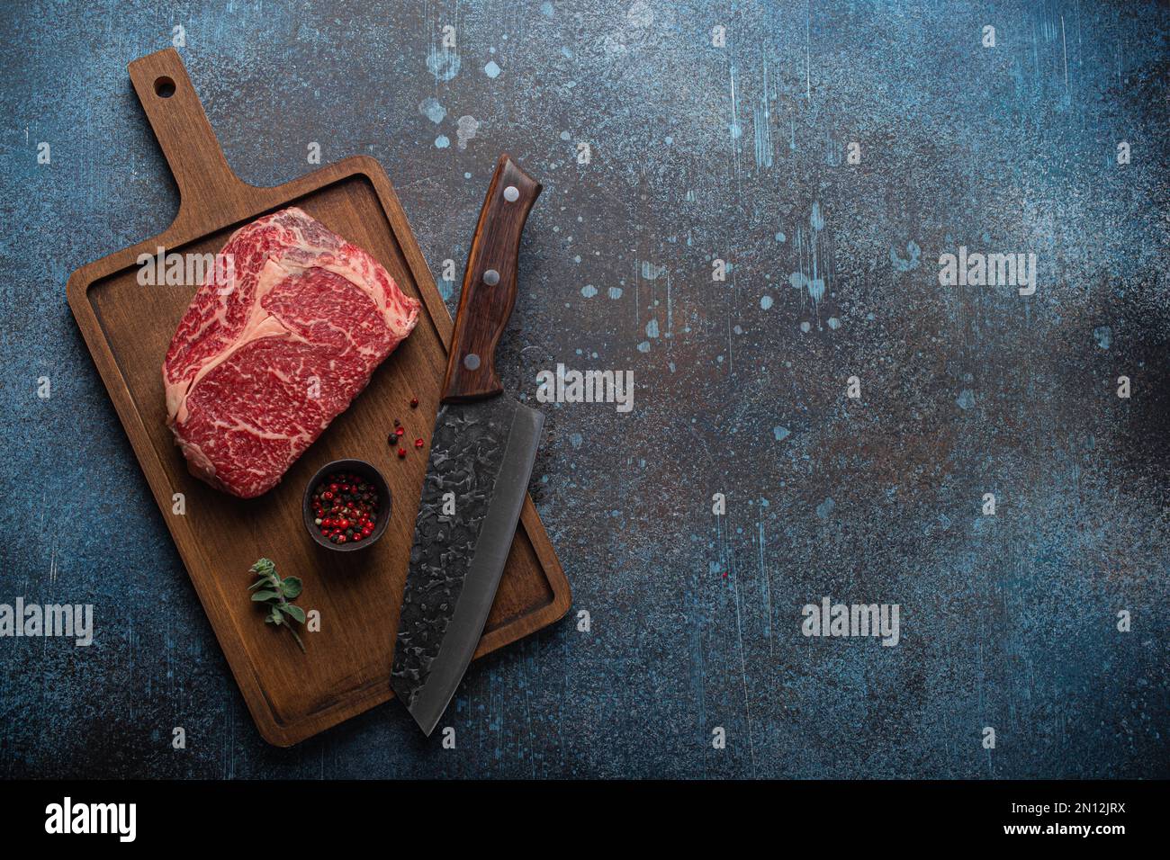 Bœuf à la viande cru bœuf marbré steak de première qualité Ribeye sur planche à découper en bois et fond de table de cuisine rustique en béton d'en haut avec un gros couteau et des épices Banque D'Images