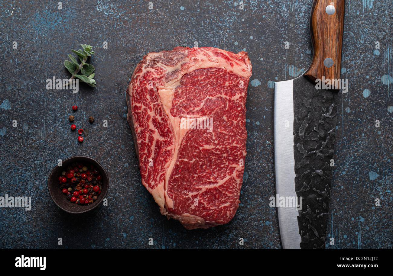 Bœuf à la viande cru bœuf marbré steak de première qualité Ribeye sur fond rustique de table de cuisine en béton d'en haut avec grand couteau et épices, concept de beefsteak Banque D'Images