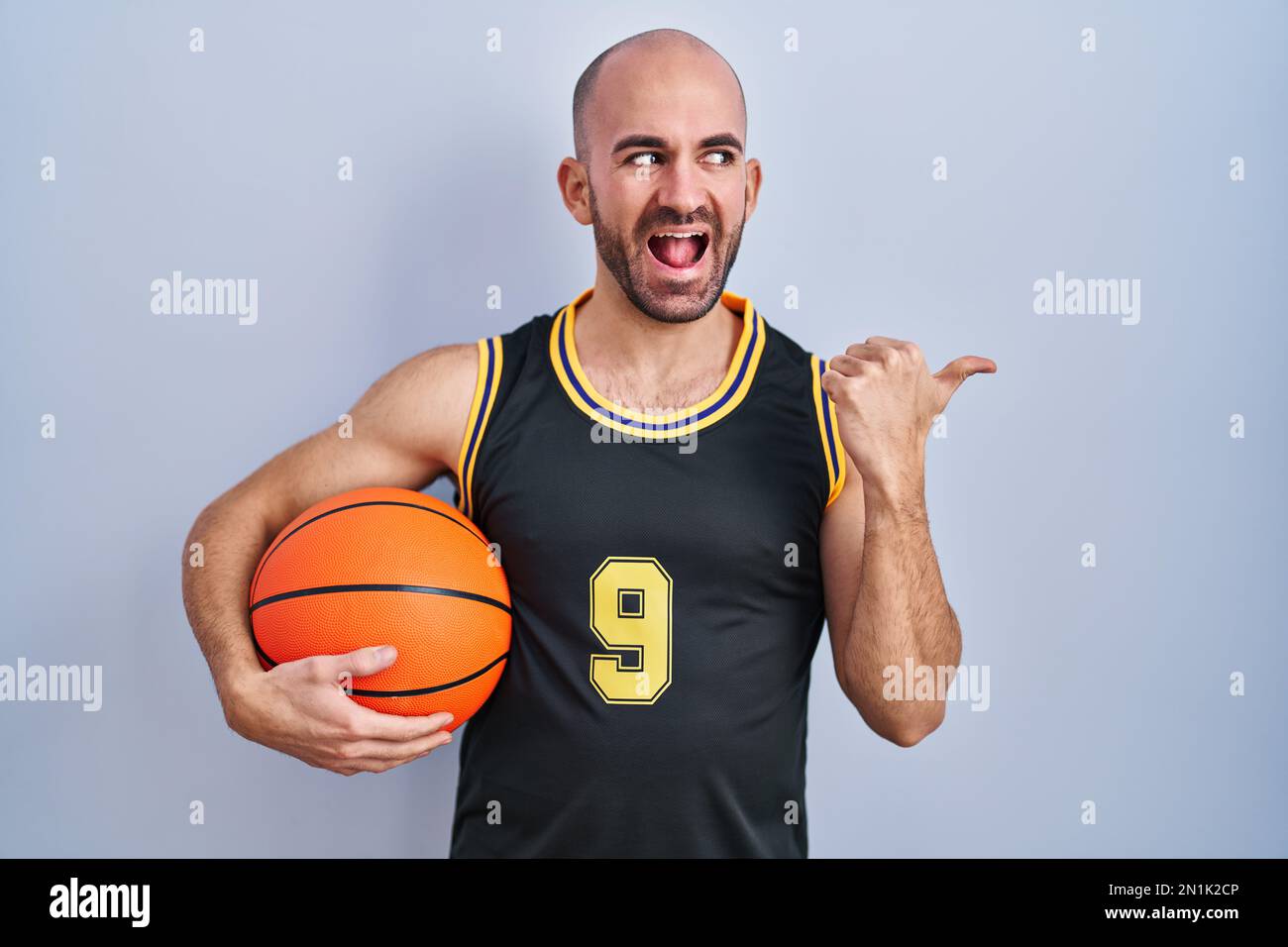 Jeune homme chauve avec barbe portant un uniforme de basket-ball tenant le  ballon souriant avec le visage heureux regardant et pointant vers le côté  avec le pouce vers le haut Photo Stock -