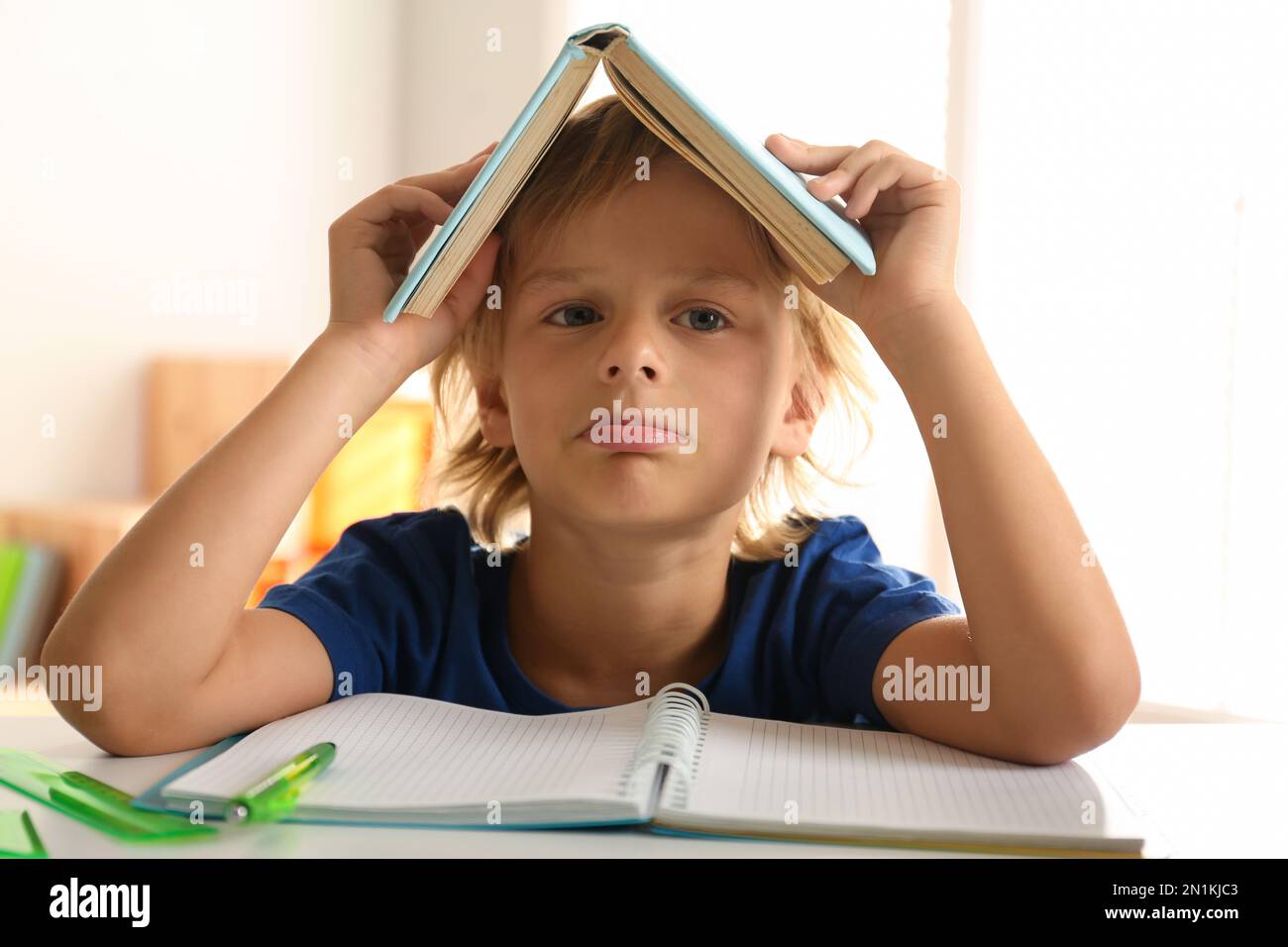 Il s'est ennuyé un petit garçon avec un livre sur sa tête, faisant ses devoirs à la table à l'intérieur Banque D'Images